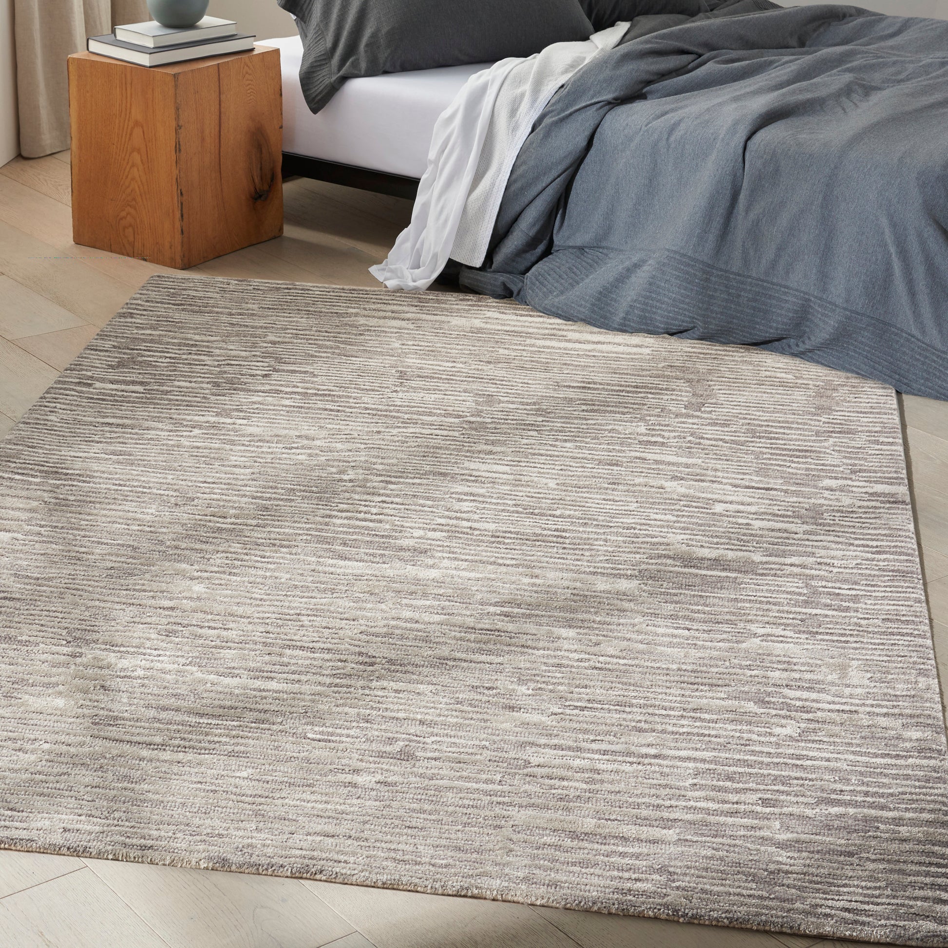 Neutral beige-gray textured area rug with subtle lines in a bedroom beside a wooden nightstand and gray bedding.