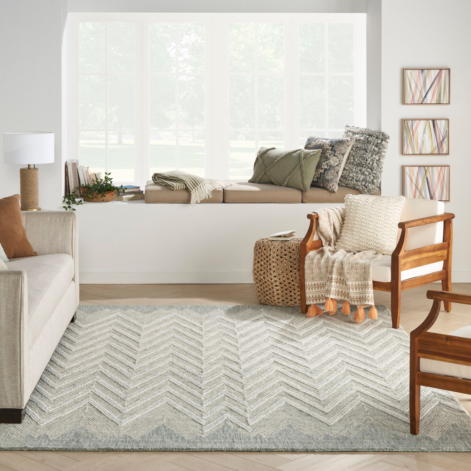 Bright living room with light gray chevron rug, beige sofa, wooden armchair, window bench, woven basket, and wall art.