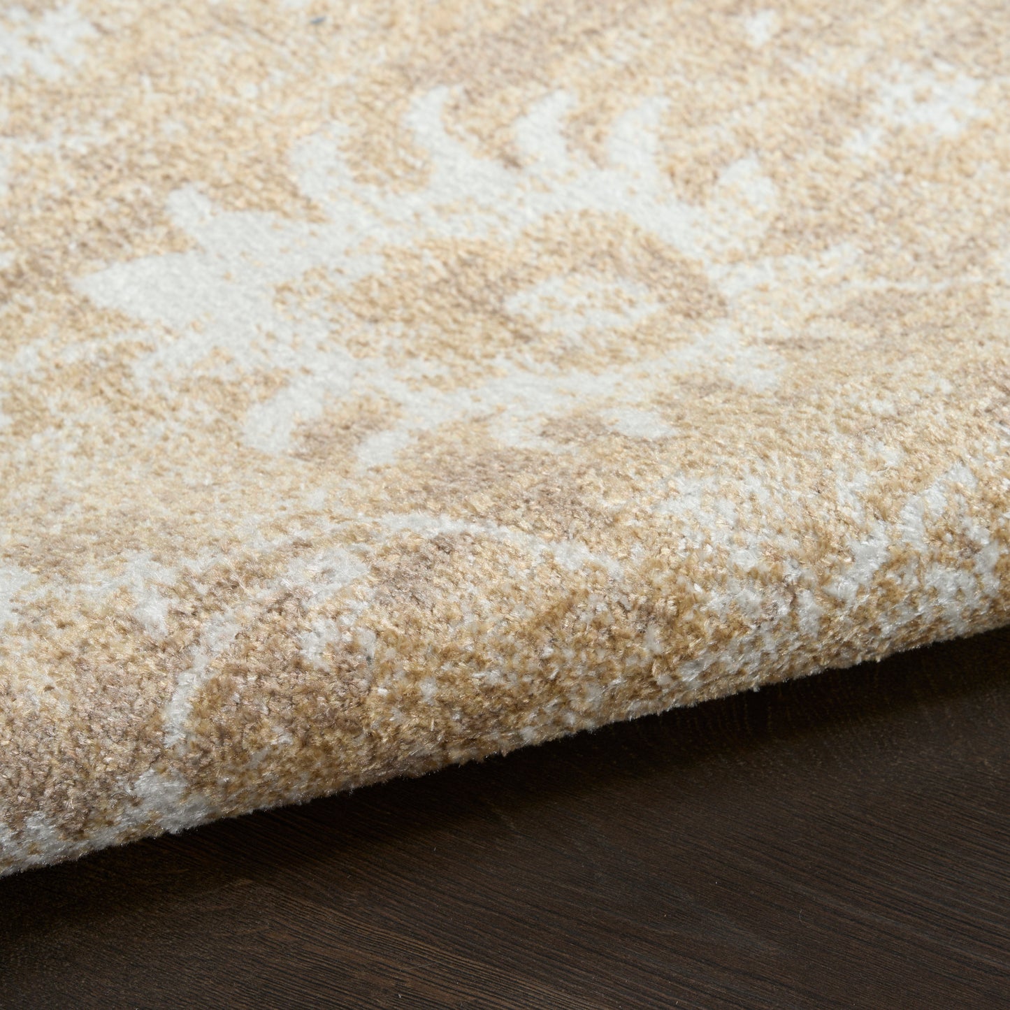 Beige area rug with cream floral pattern, soft plush pile, edge folded on dark wood floor.