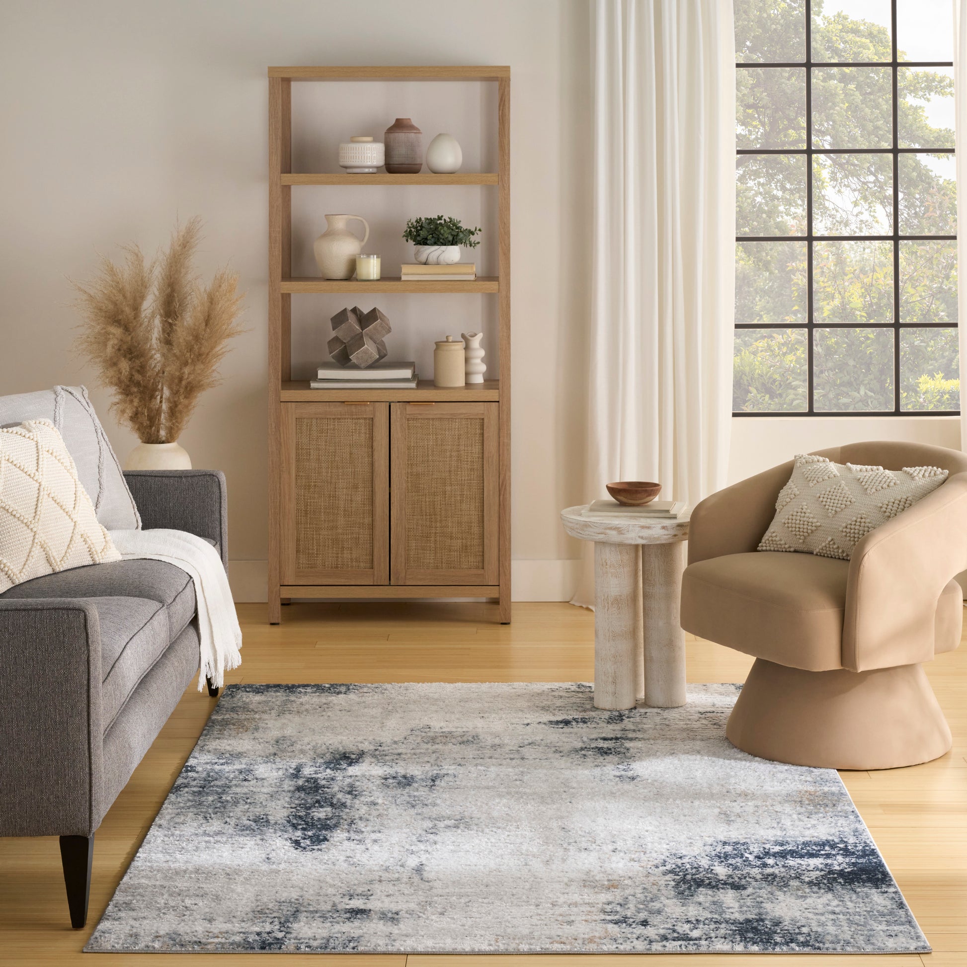 Living room with natural wood open shelf, woven cabinet doors, gray sofa, beige curved chair, blue-gray abstract rug, and large grid window.