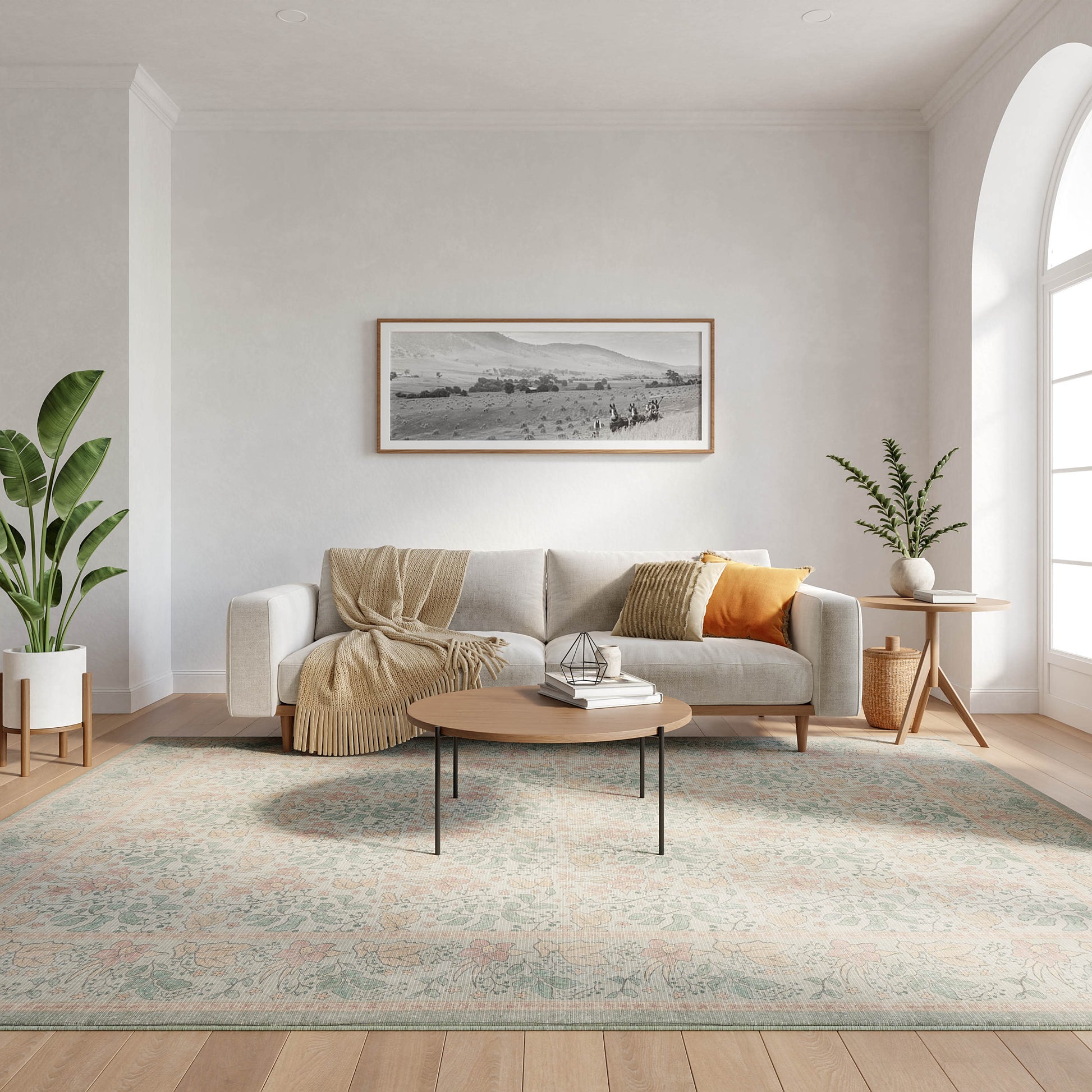Neutral living room: light gray sofa, beige throw, orange pillow, round wood coffee table, pastel floral rug, plants.