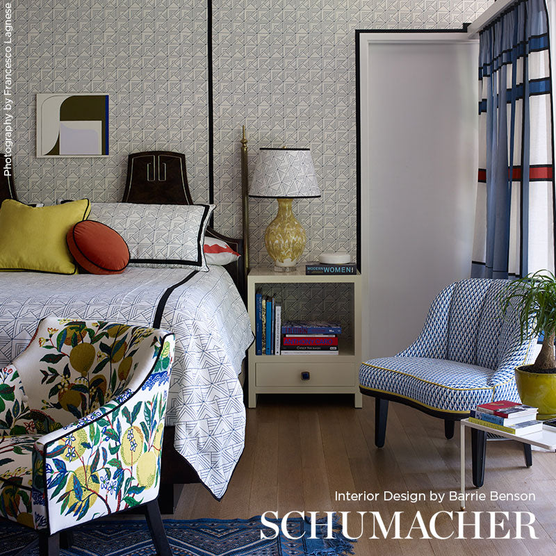 Chic bedroom with geometric wallpaper, white quilt bed, yellow cushions, blue armchair, lamp on nightstand, striped curtains.
