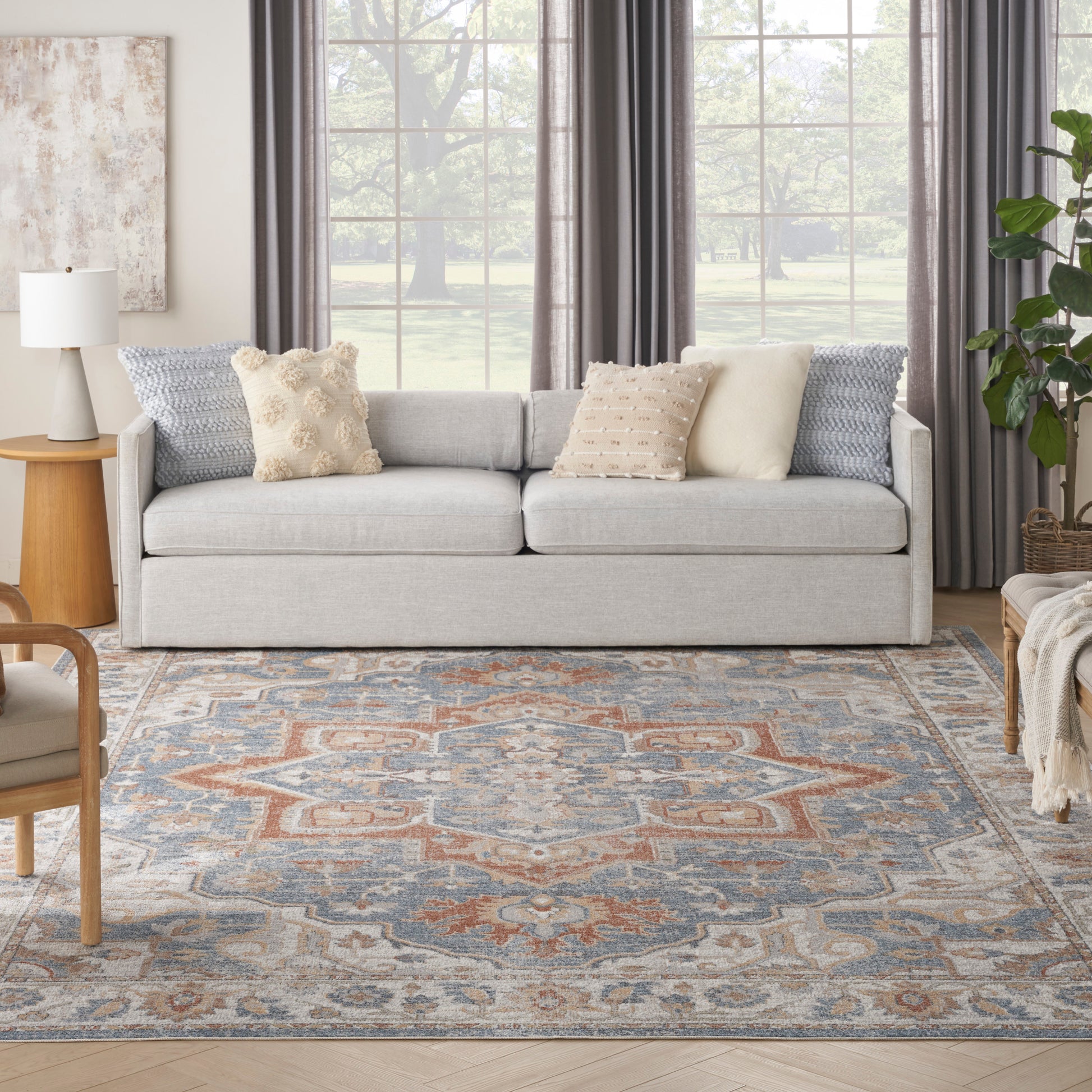 Patterned blue rug on the floor with a light gray sofa and textured beige and blue cushions, a wooden side table, and a large window with gray drapes.