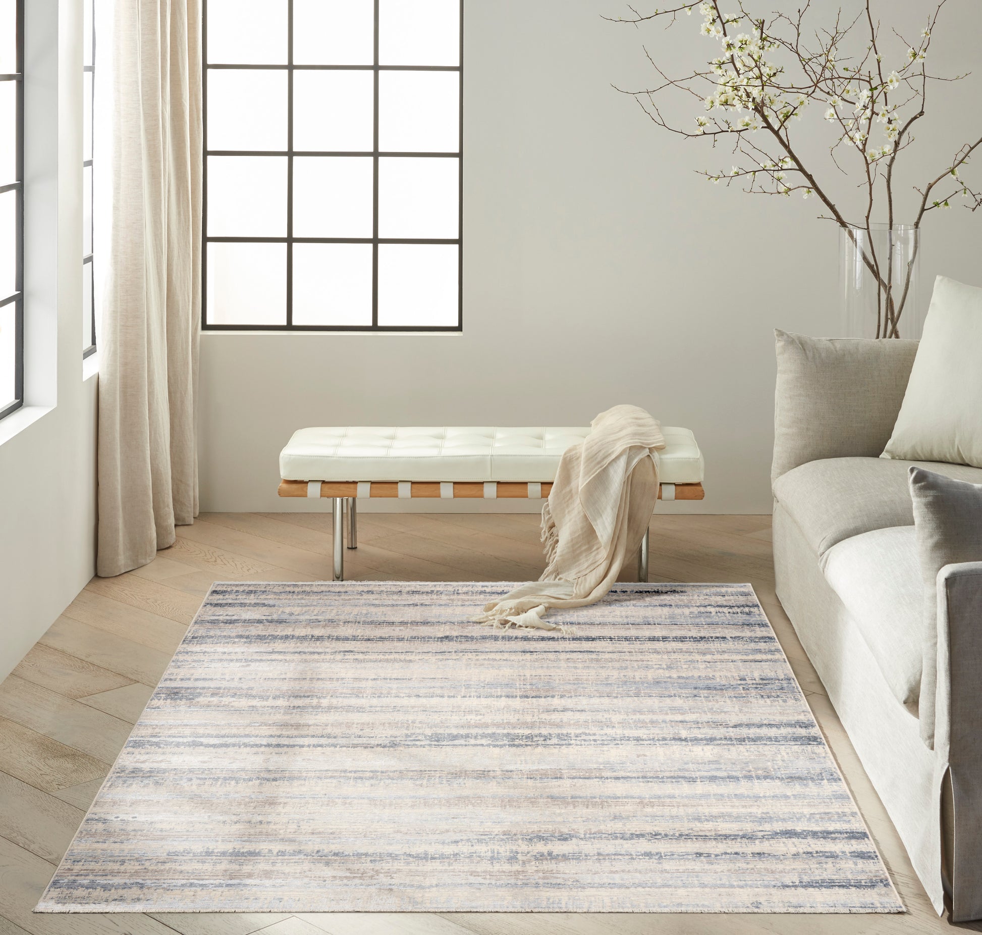 Neutral living room with gradient blue-gray rug, white tufted bench with wood slats & chrome legs, beige throw, gray sofa.