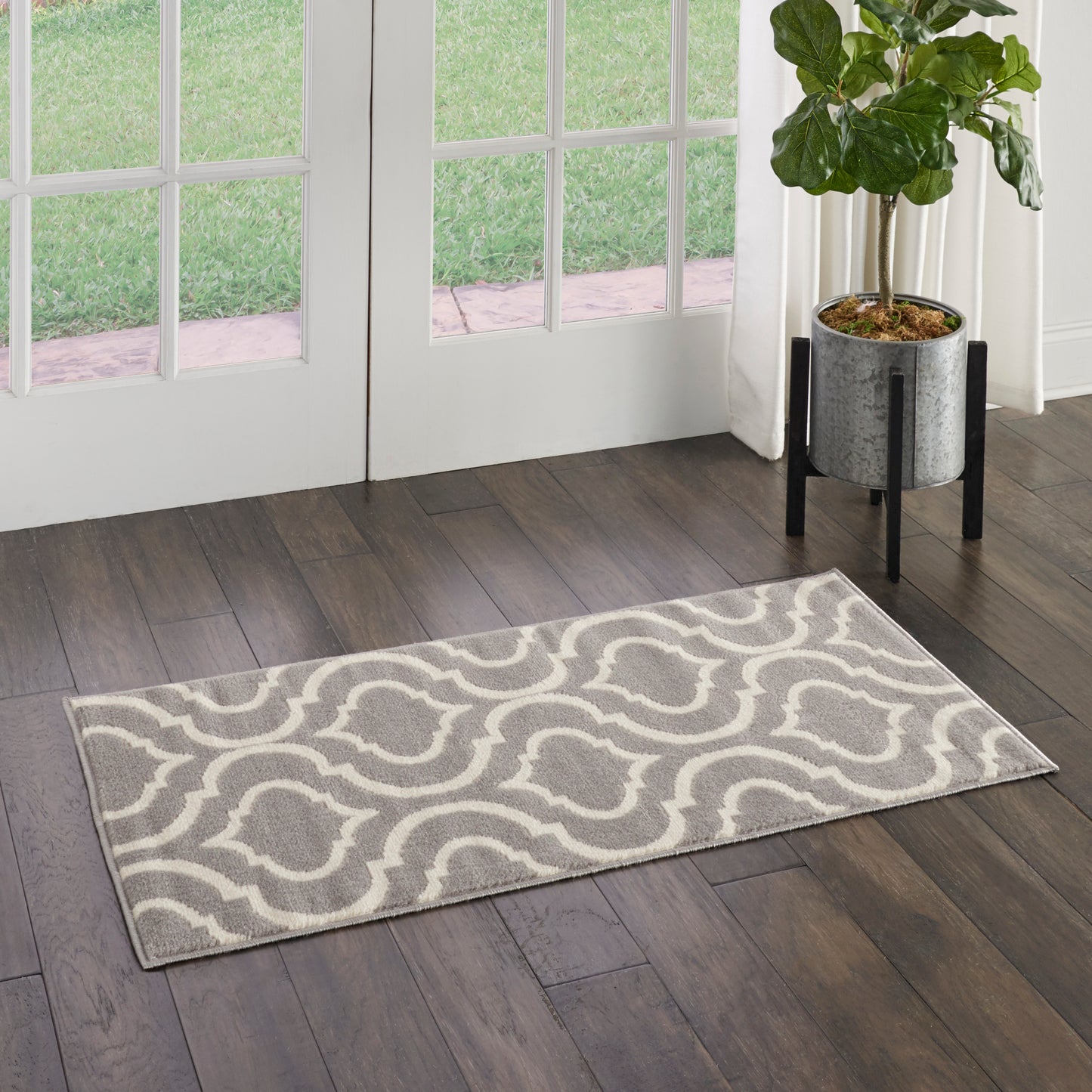 Grey rectangular rug with white wavy geometric pattern on dark wood floor beside glass doors, potted plant nearby.