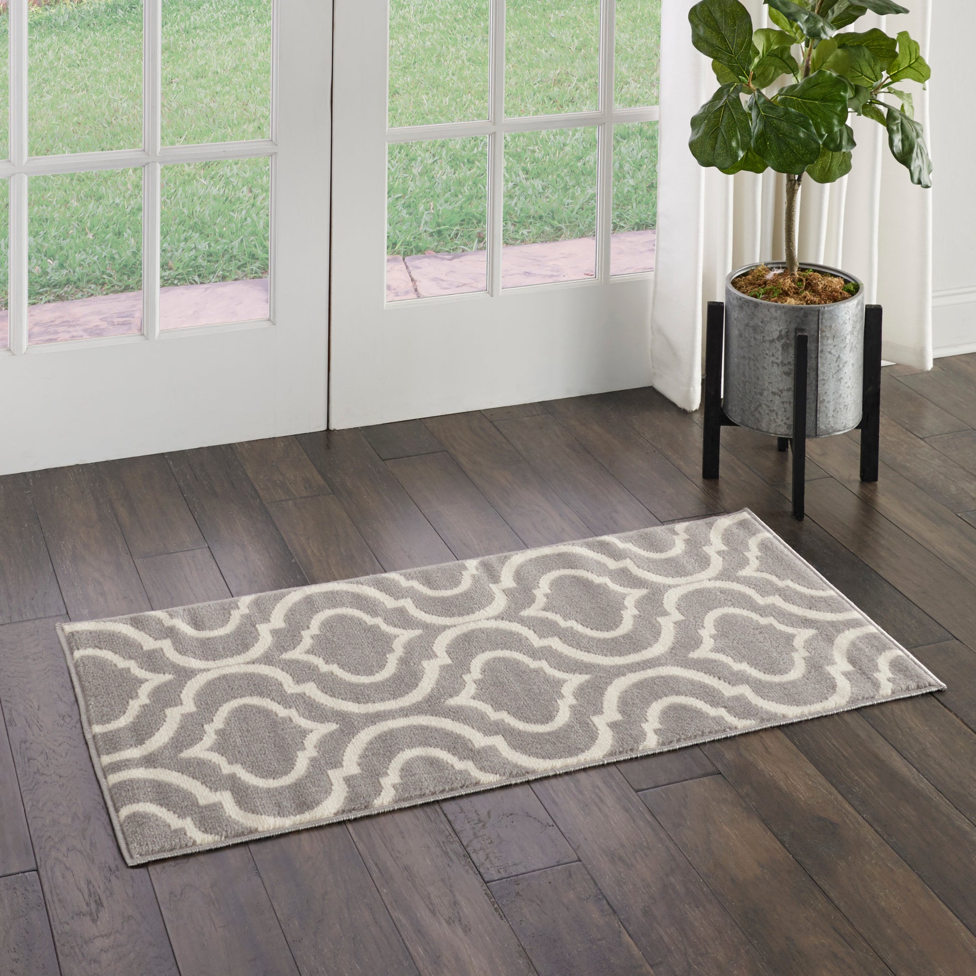 Grey rectangular rug with white wavy geometric pattern on dark wood floor beside glass doors, potted plant nearby.