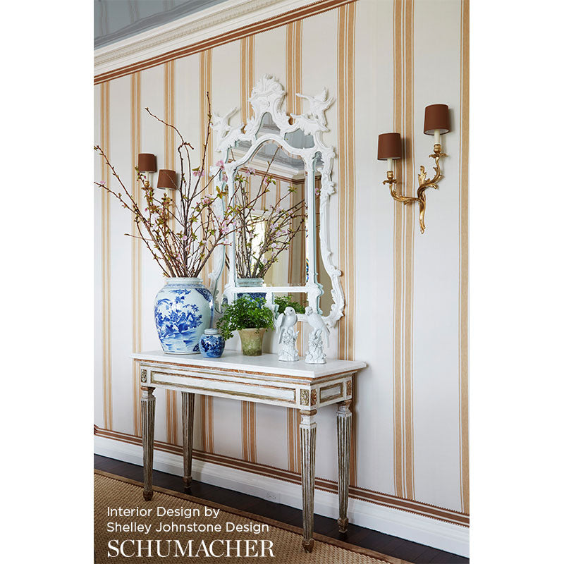 Distressed white console with gold trim, ornate mirror, blue-and-white vase with branches, striped wallpaper, brass sconces.