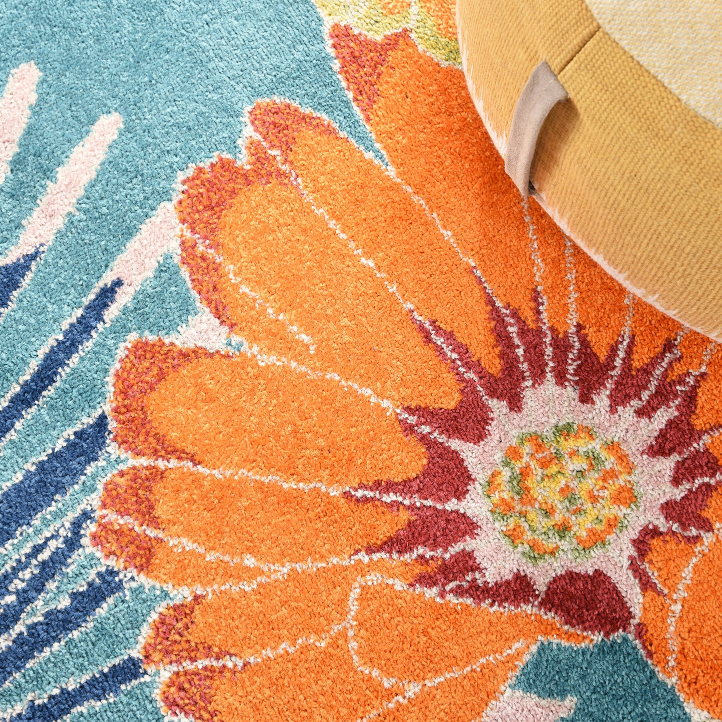 Close-up tufted rug with a large orange flower on teal blue background; beige straw hat at edge.
