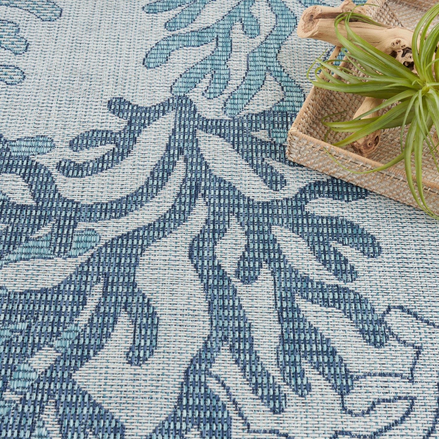 Blue & light-blue woven rug with leafy botanical pattern; natural wicker basket with a green plant sits at the edge.