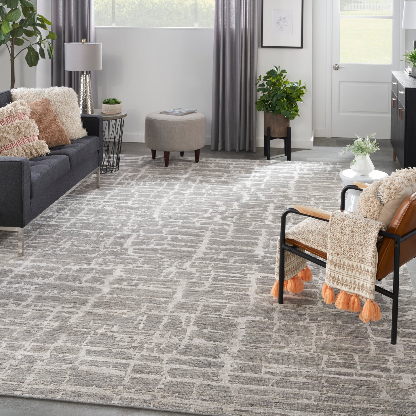Cozy modern living room: gray sofa, beige round ottoman, metal-frame chair with orange tassel throw, geometric gray rug