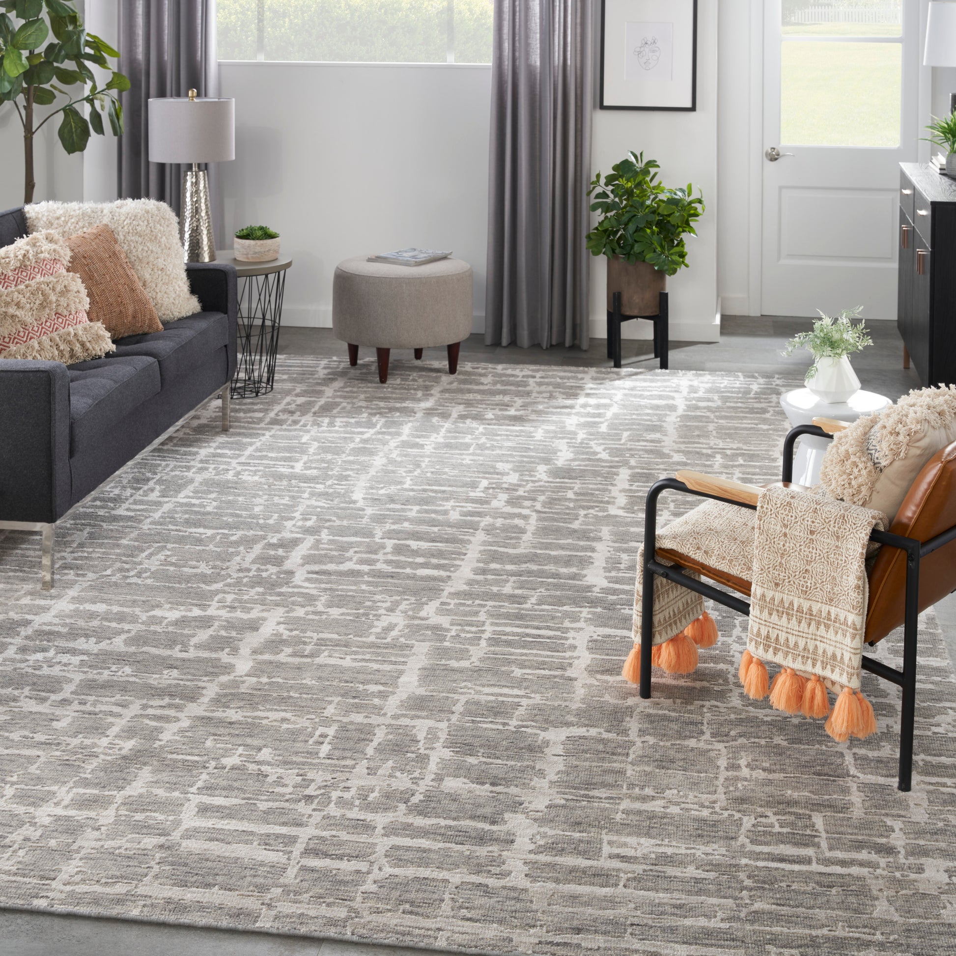 Cozy modern living room: gray sofa, beige round ottoman, metal-frame chair with orange tassel throw, geometric gray rug