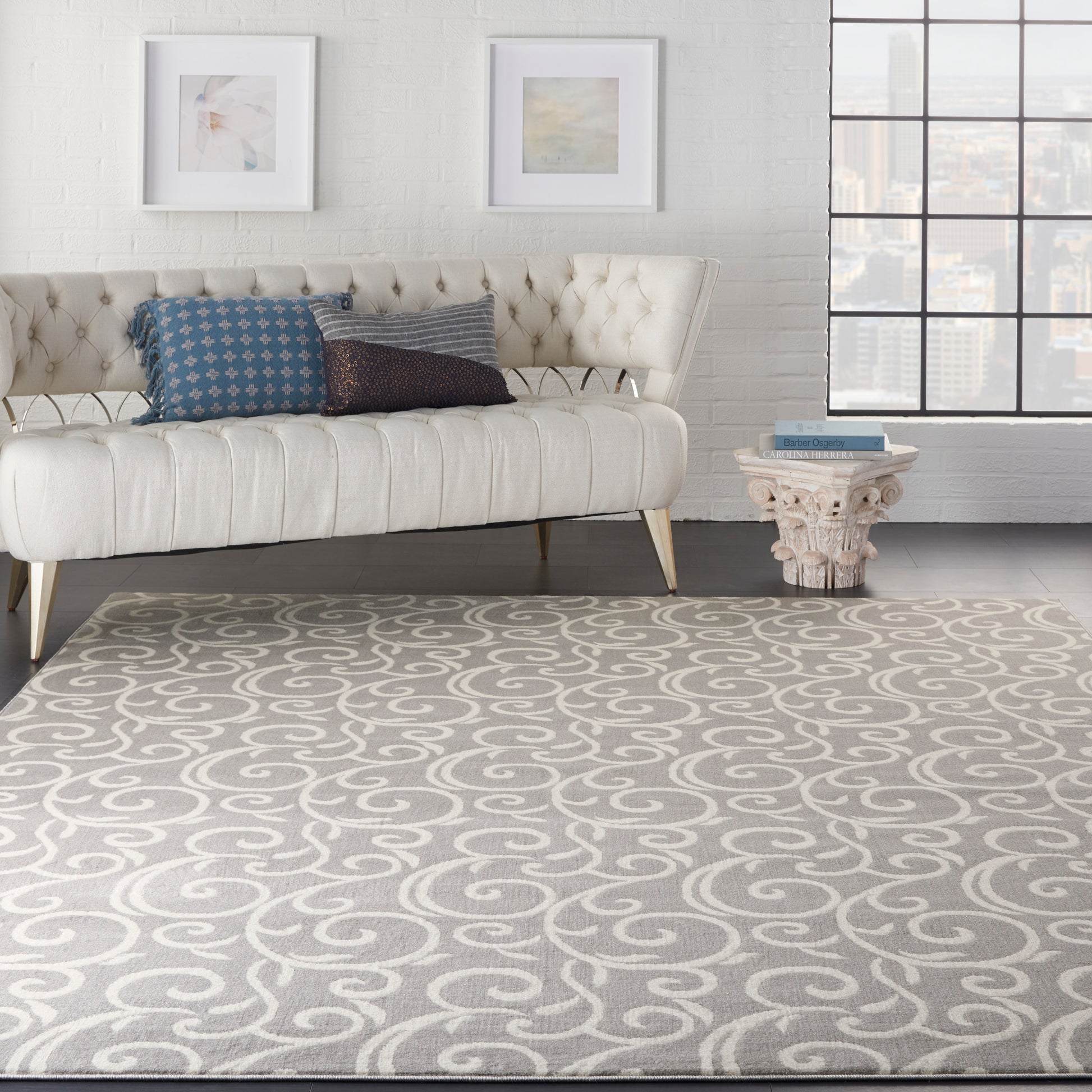 Gray swirl rug on a light floor in a living room, with a cream tufted sofa and blue pillows, carved round side table, white brick wall and a city view through the window.
