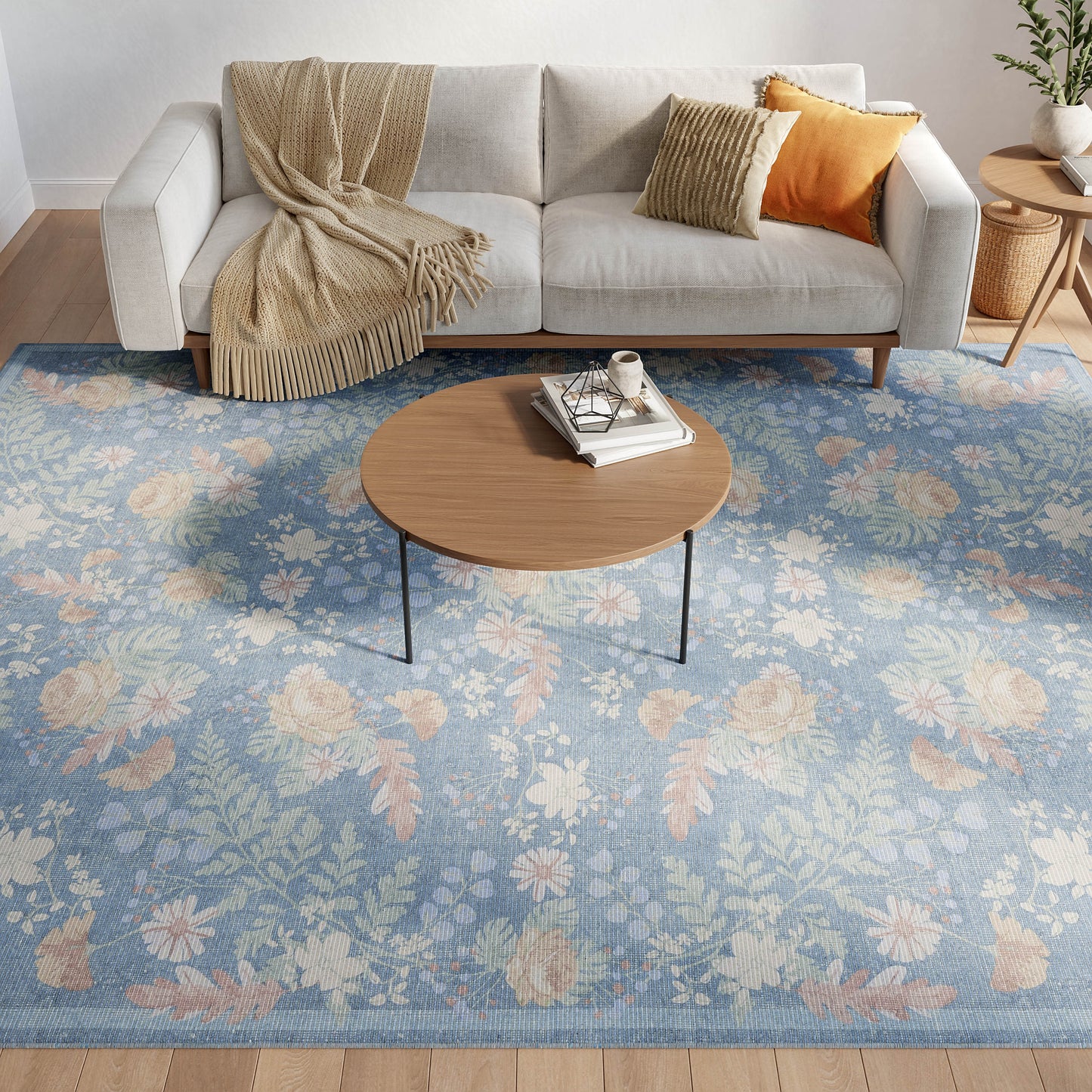 Living room scene: round wood coffee table with slim metal legs, blue floral area rug, gray sofa with beige fringed throw and orange cushions.