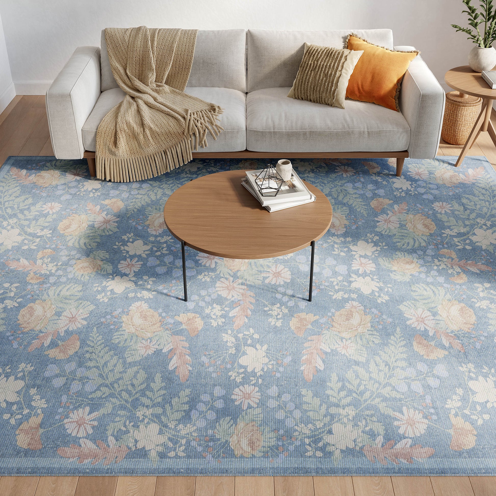 Living room scene: round wood coffee table with slim metal legs, blue floral area rug, gray sofa with beige fringed throw and orange cushions.