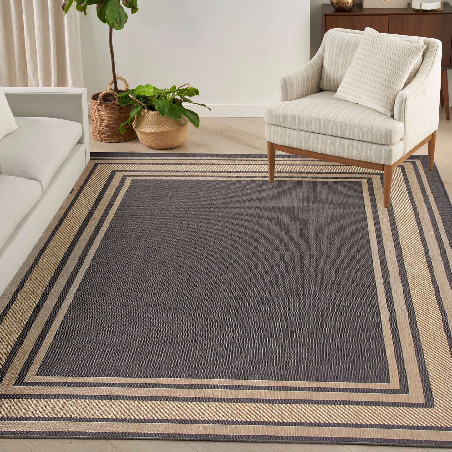 Charcoal gray rectangular area rug (10' x 14') with layered beige borders in a modern living room, beside a cream striped armchair and plants.