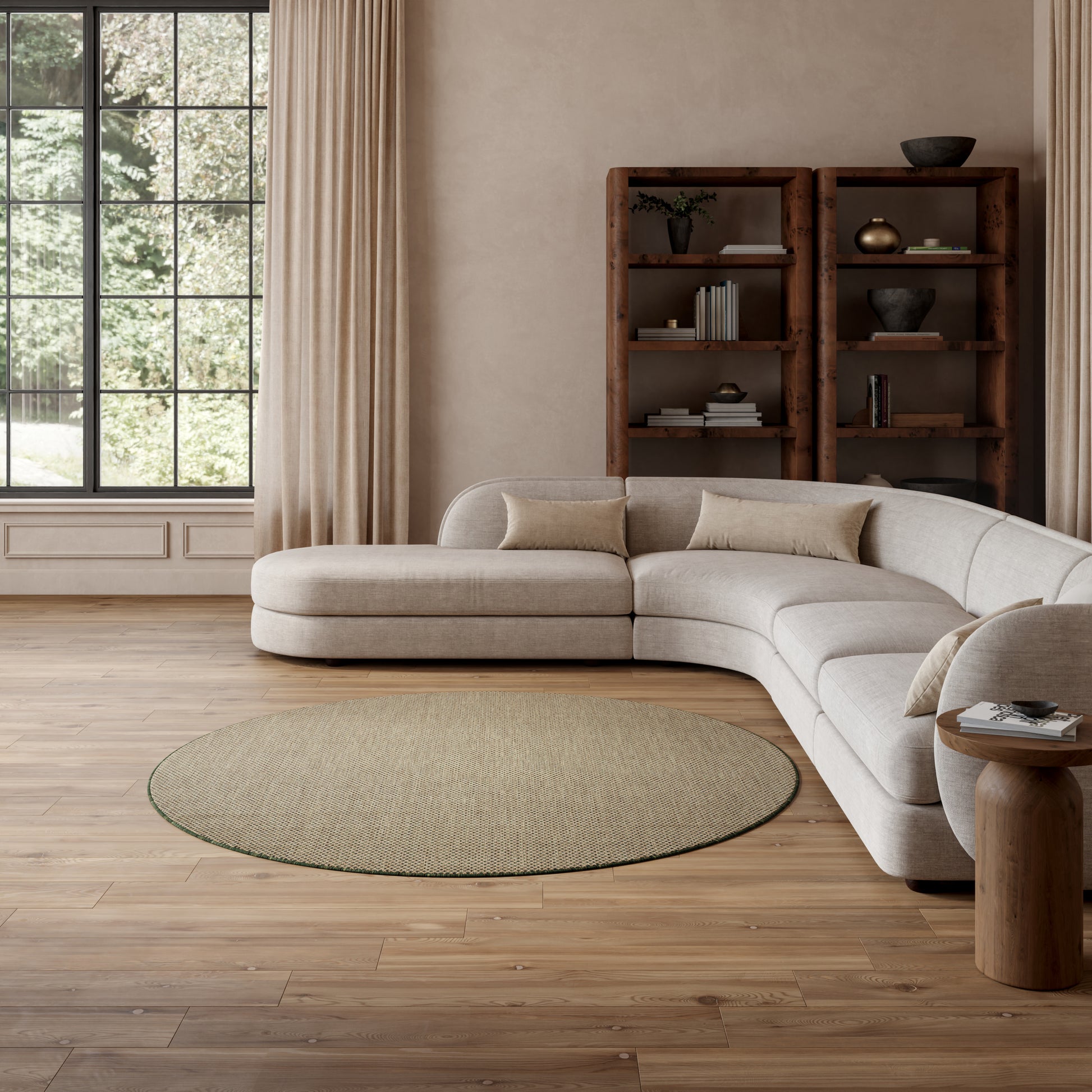 Beige L-shaped sectional sofa in a bright living room with a round natural fiber rug, wood bookshelf, and tall windows.