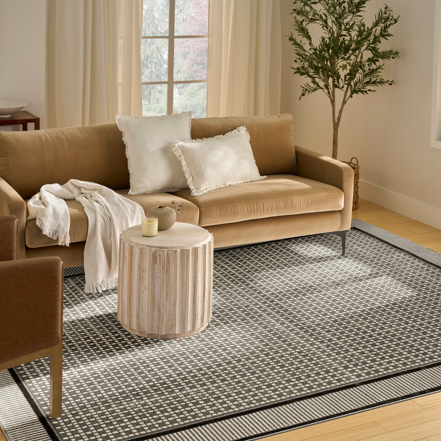 Black and white rug on a beige living room floor, with a beige fabric sofa featuring white fringe pillows and a cream throw, a round grooved wood side table, and a lit candle.