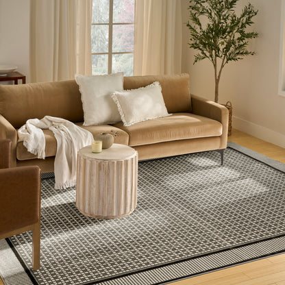 Black and white rug on a beige living room floor, with a beige fabric sofa featuring white fringe pillows and a cream throw, a round grooved wood side table, and a lit candle.