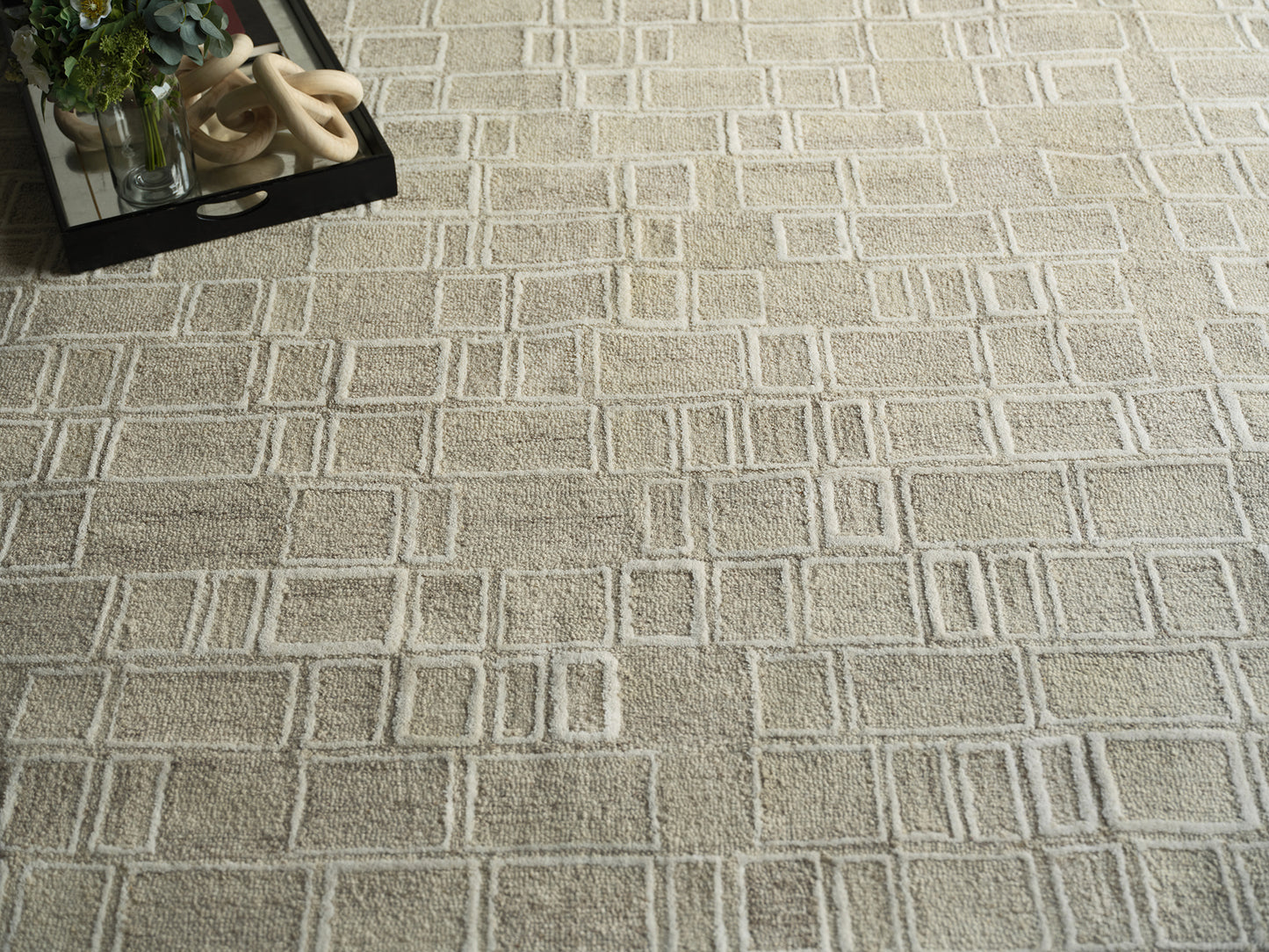 Beige geometric area rug with a raised square grid; top-left shows a black tray, plant, and beige ring sculpture.