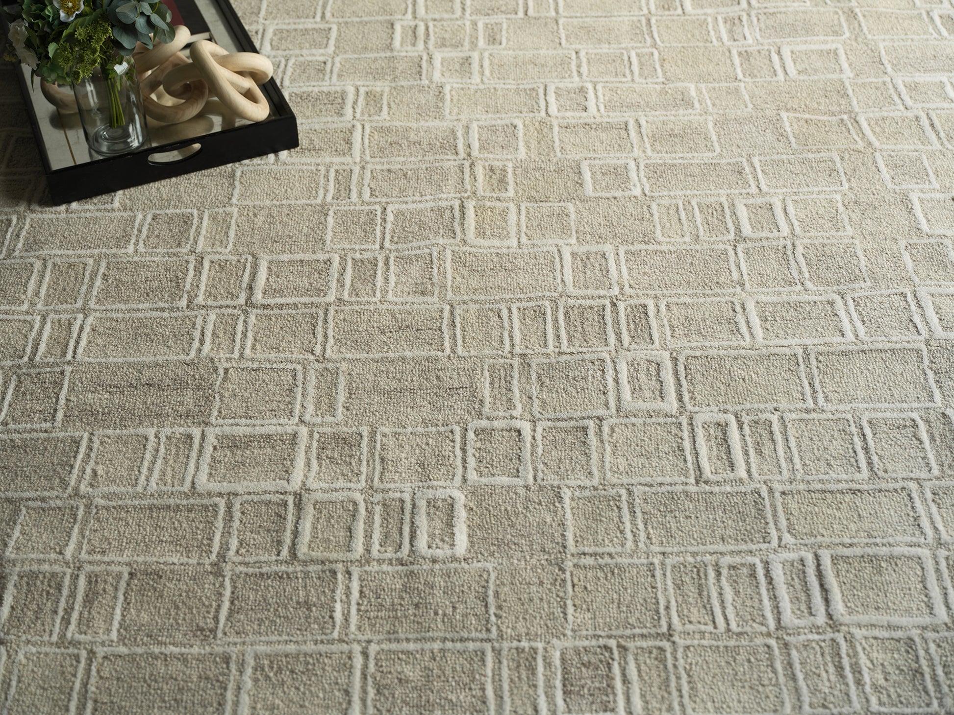 Beige geometric area rug with a raised square grid; top-left shows a black tray, plant, and beige ring sculpture.