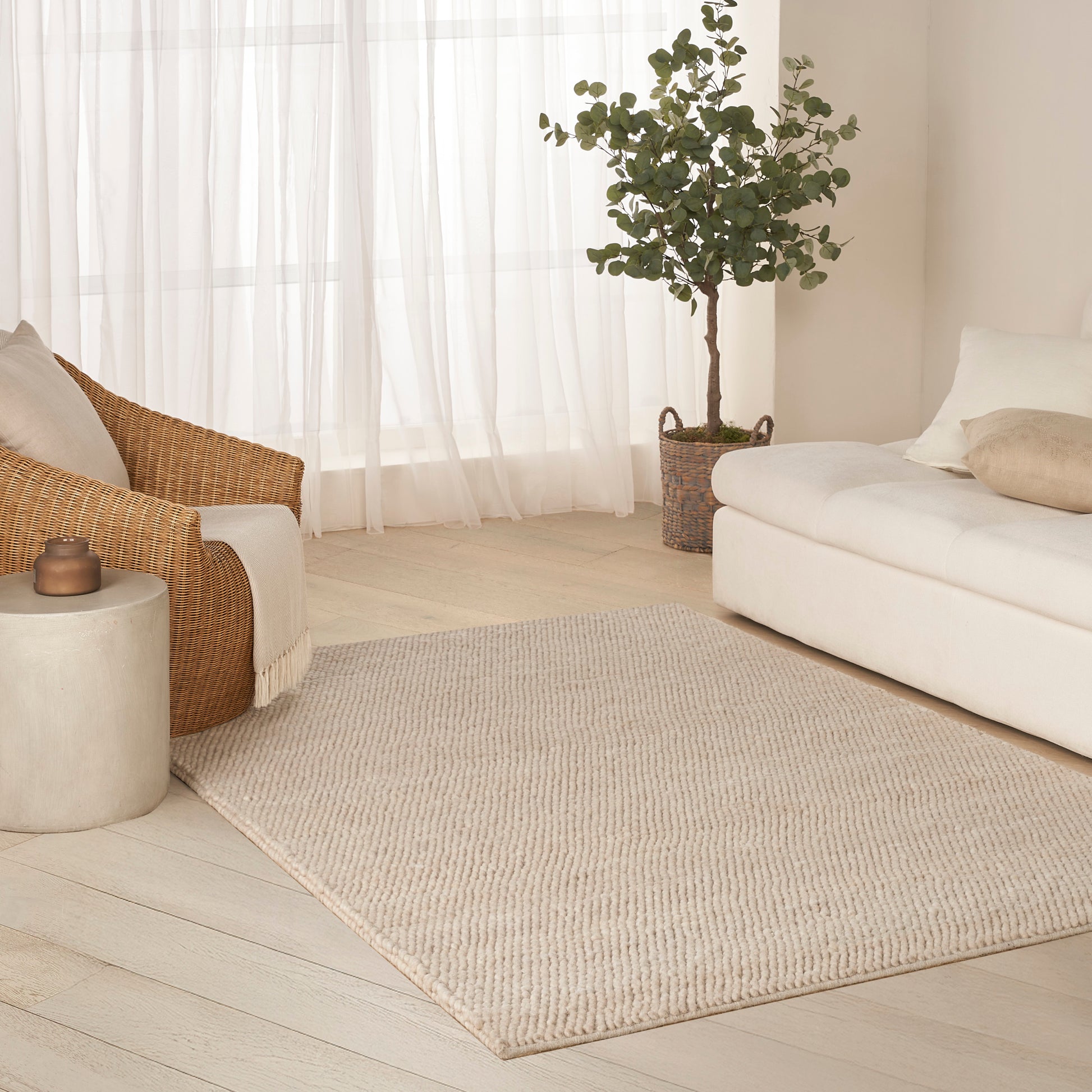 Beige jute rug on light wood floor with cream sofa, wicker chair, sheer curtains, and a potted plant.