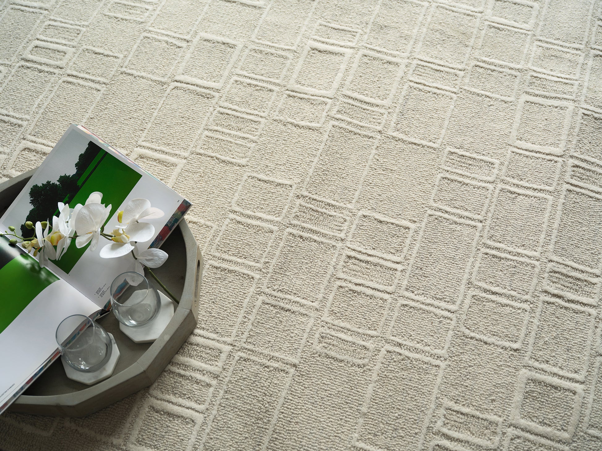 Beige geometric textured rug with a round tray holding a magazine spread, white orchid bouquet, and two glass candle holders.