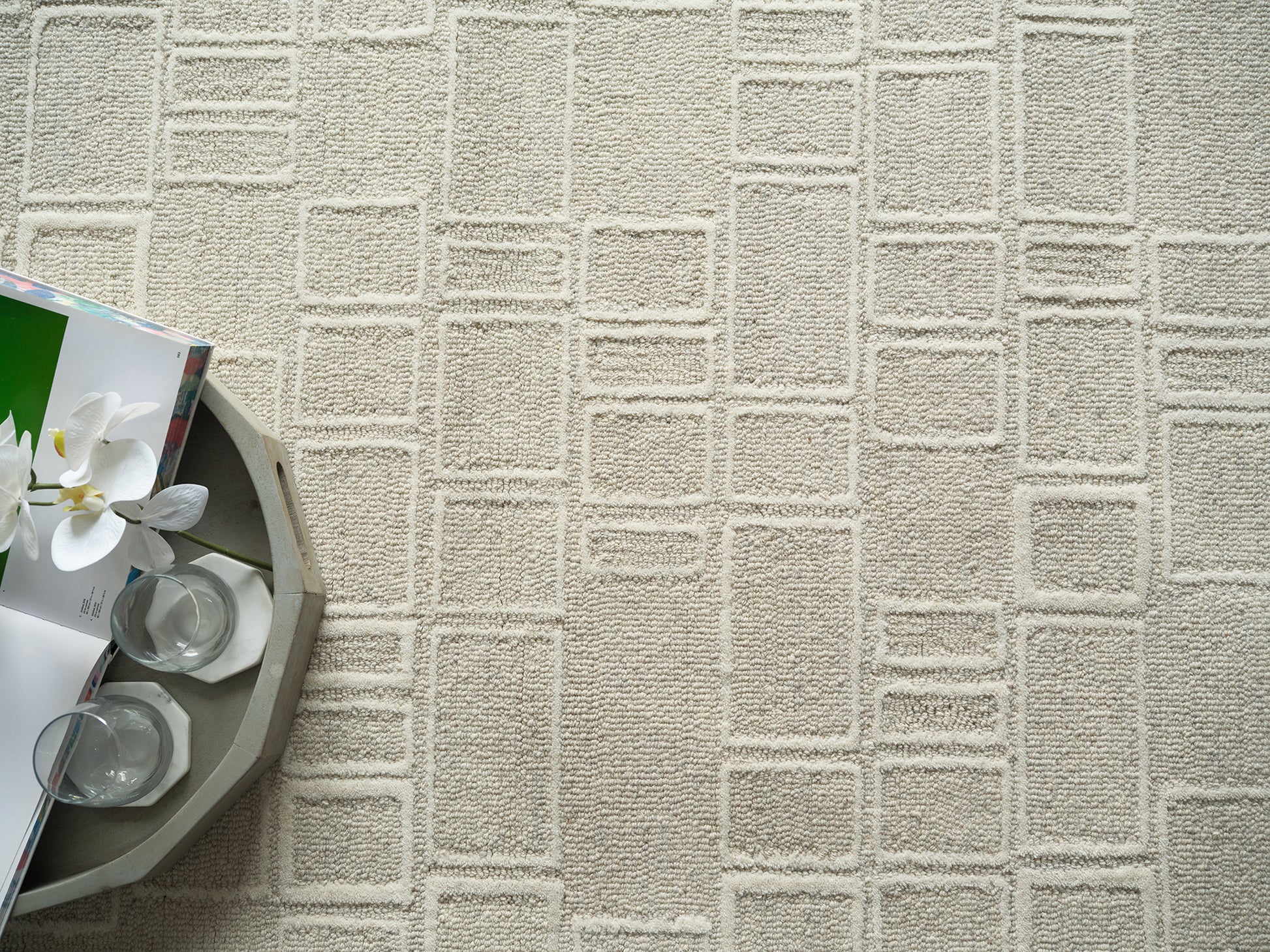 Beige textured rug with a square-grid pattern; a round tray holds three glass candle holders and a magazine featuring a white orchid.