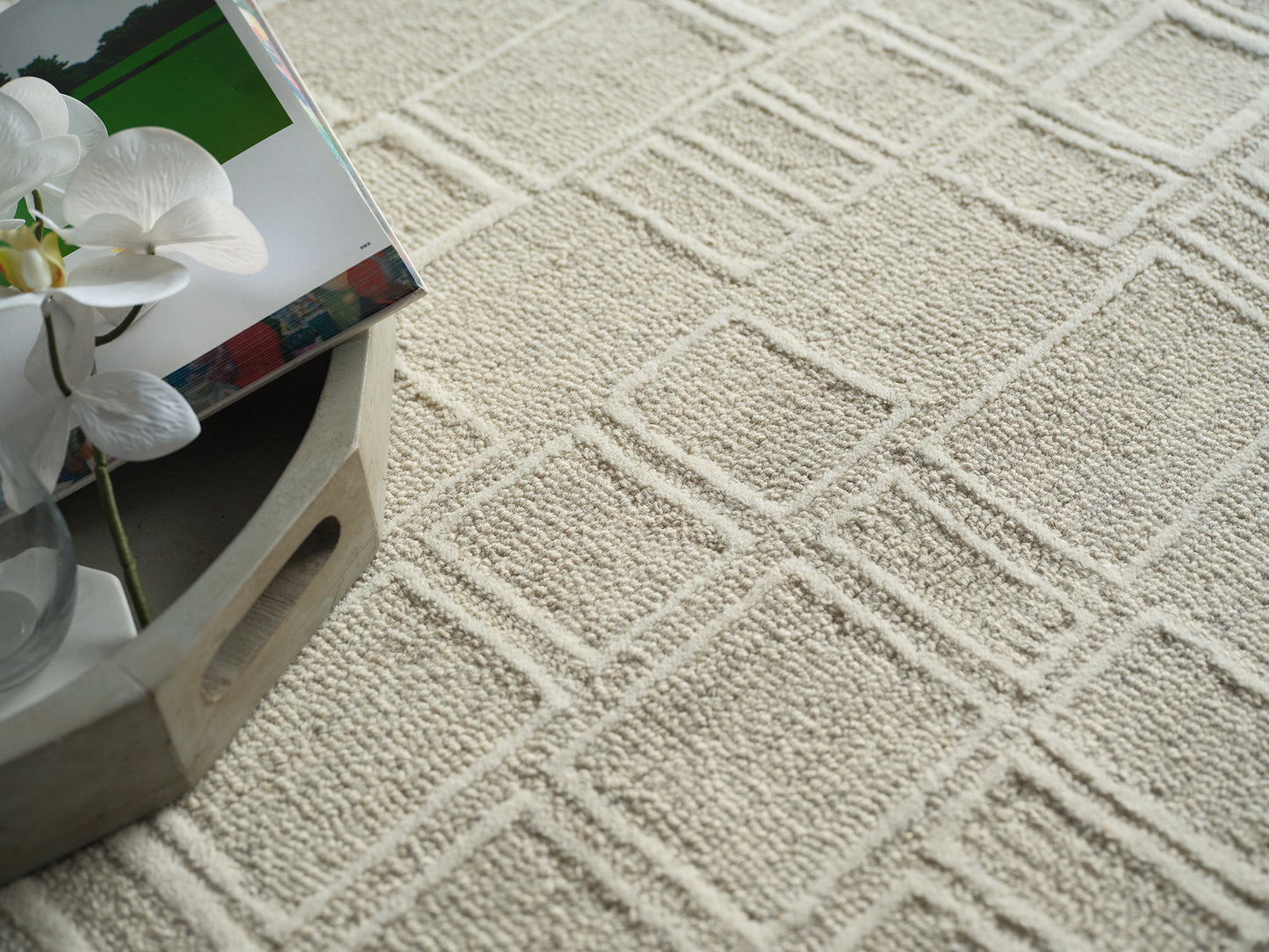 Beige textured rug with a square-diamond pattern; round wooden tray holds a white orchid & a glossy magazine.