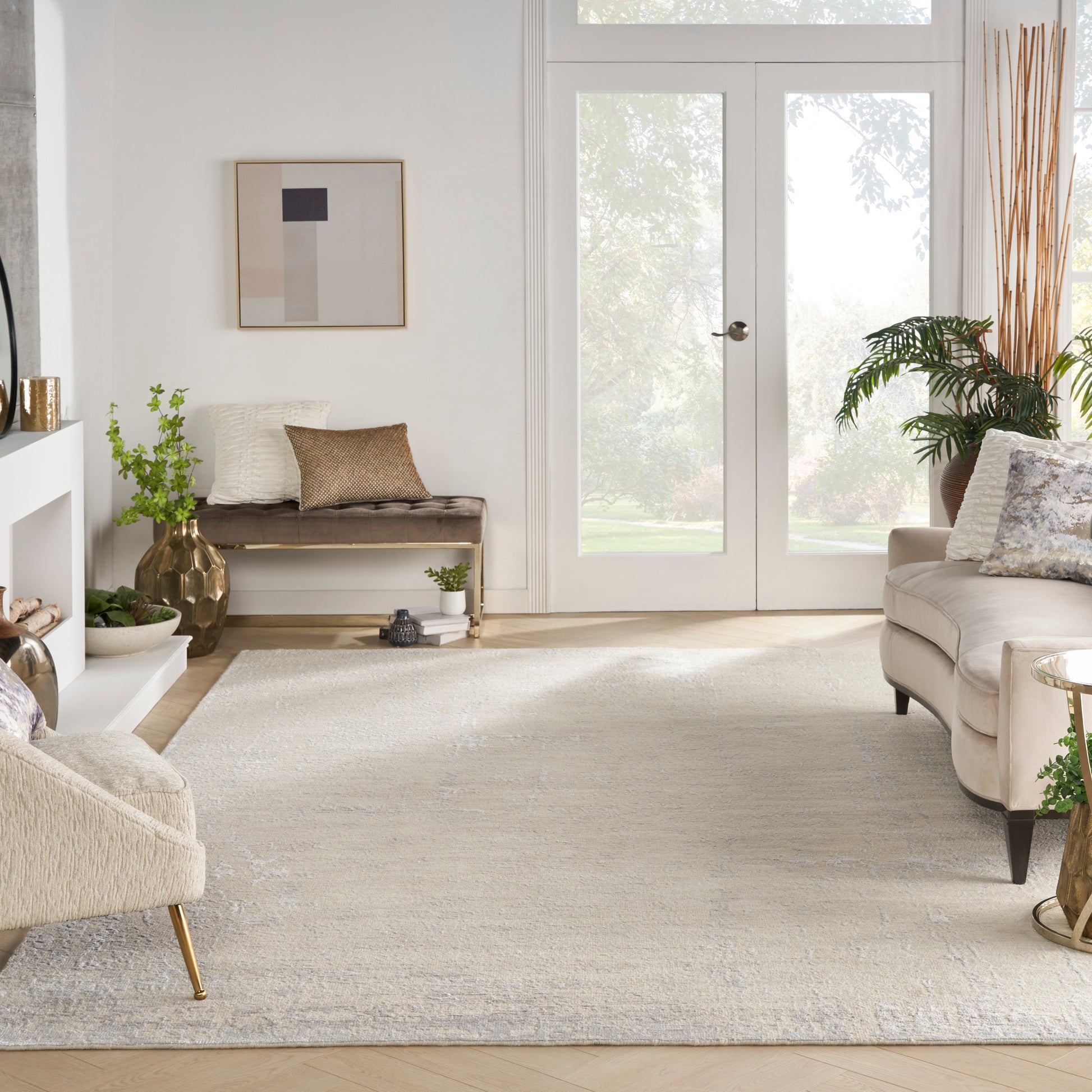 Bright neutral living room with cream sofa, beige bench, pale patterned rug, glass doors, wood floor, abstract wall art, and plants.