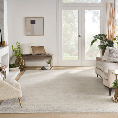 Bright neutral living room with cream sofa, beige bench, pale patterned rug, glass doors, wood floor, abstract wall art, and plants.