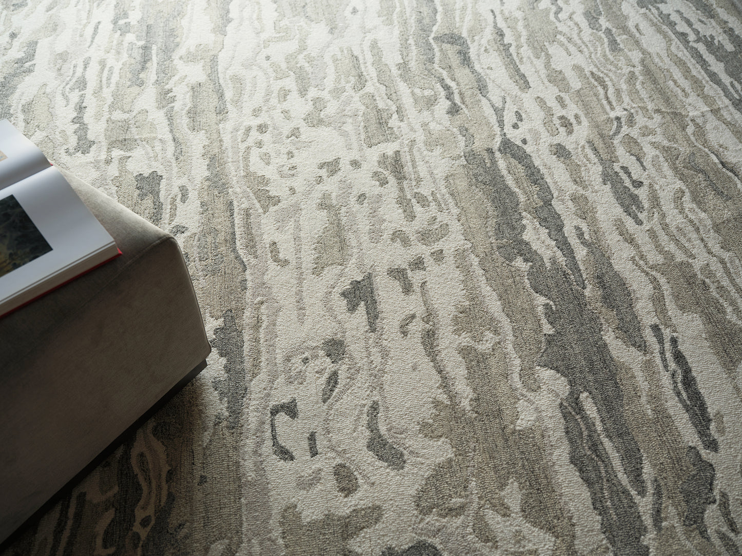 Abstract gray and beige rug with a wavy pattern beside a light tan ottoman and an open magazine.