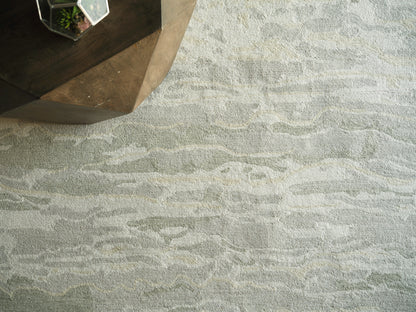 Modern abstract indoor area rug in light gray and ivory with a soft, textured pile; 6' x 9' size; corner of dark wood furniture visible.