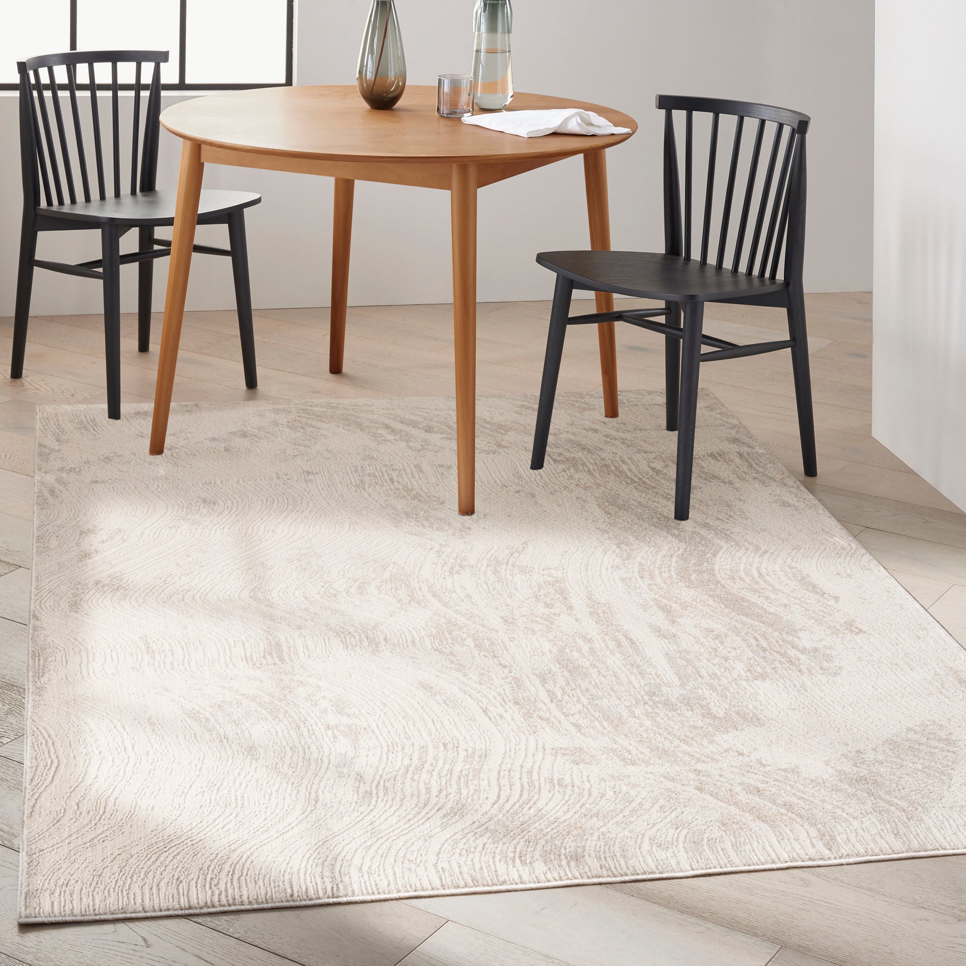 Round natural wood dining table with tapered legs, paired with black spindle-back chairs on a light beige rug.