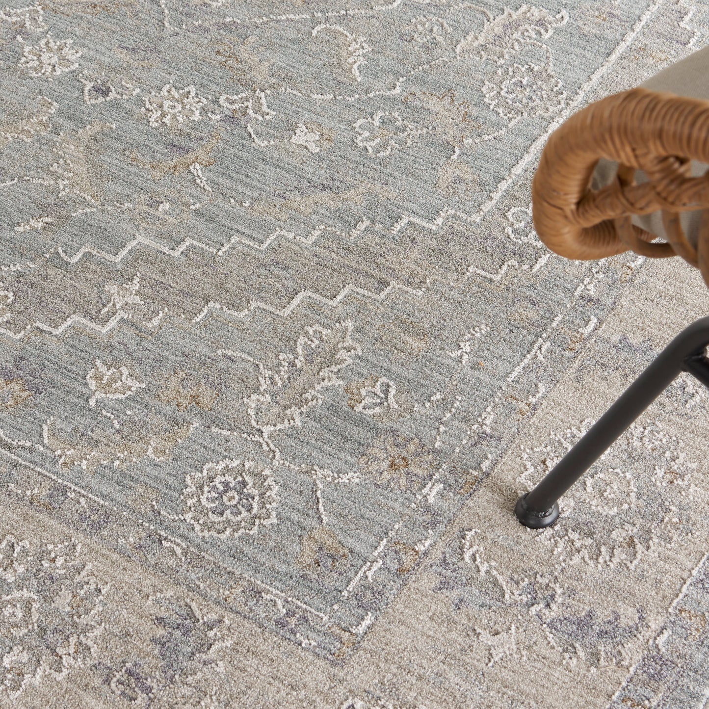 Blue-gray oriental rug with ivory floral motifs & zigzag borders; wicker chair leg visible on the right.
