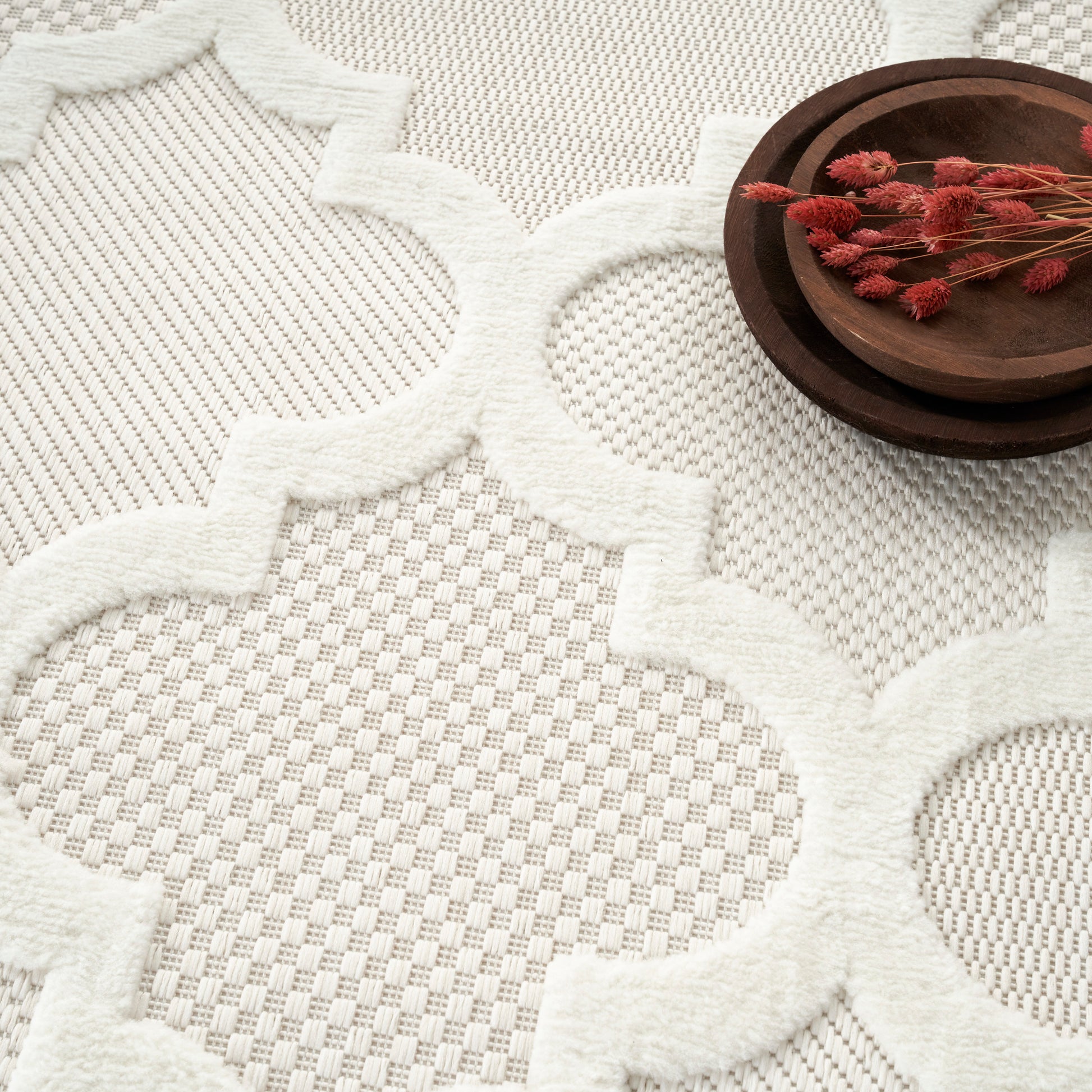 White shag rug with raised quatrefoil lattice and plush edges; dark wood bowl with red dried flowers.
