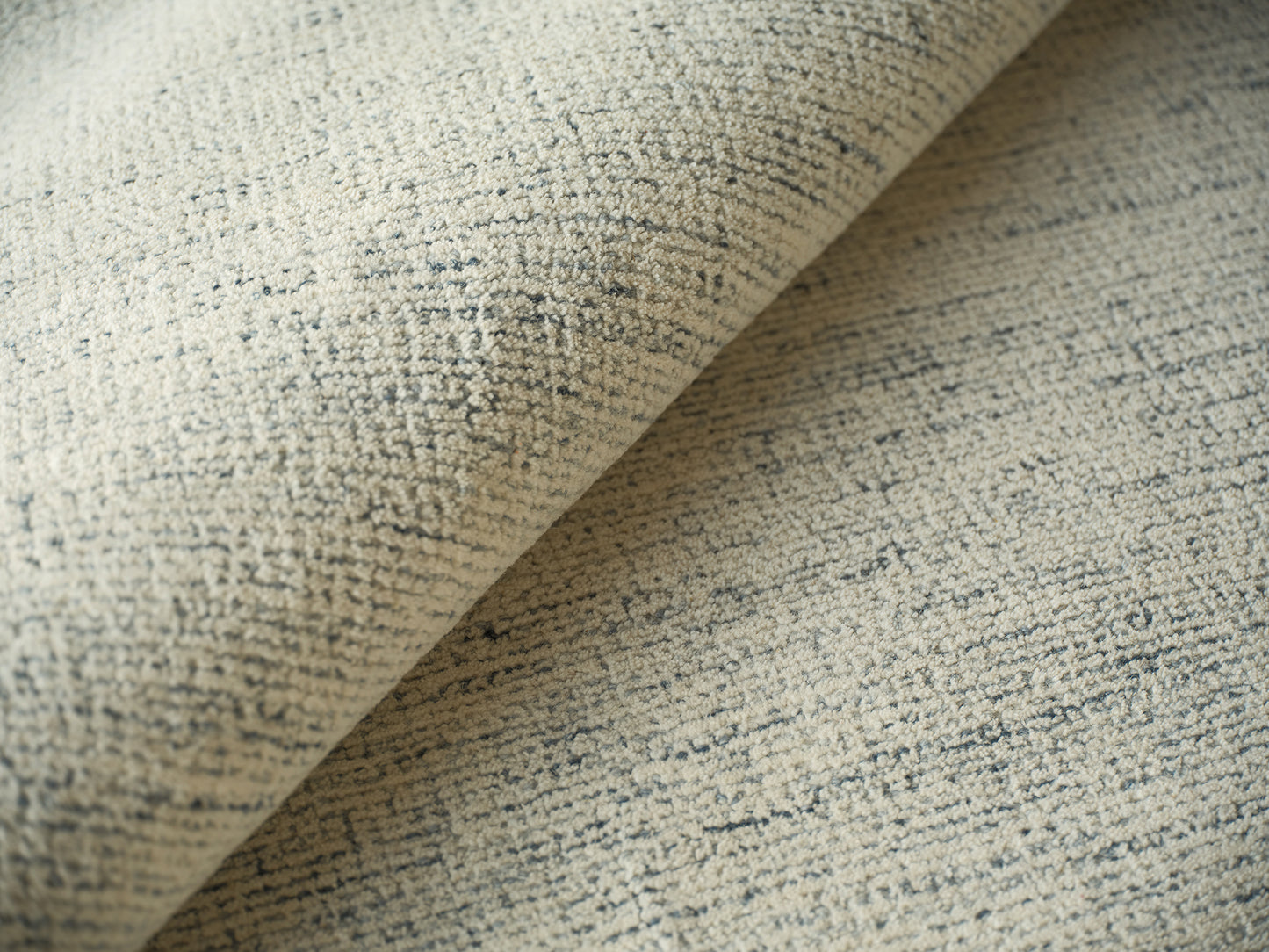 Beige textured rug with subtle blue-gray speckled pattern, rolled edge visible.