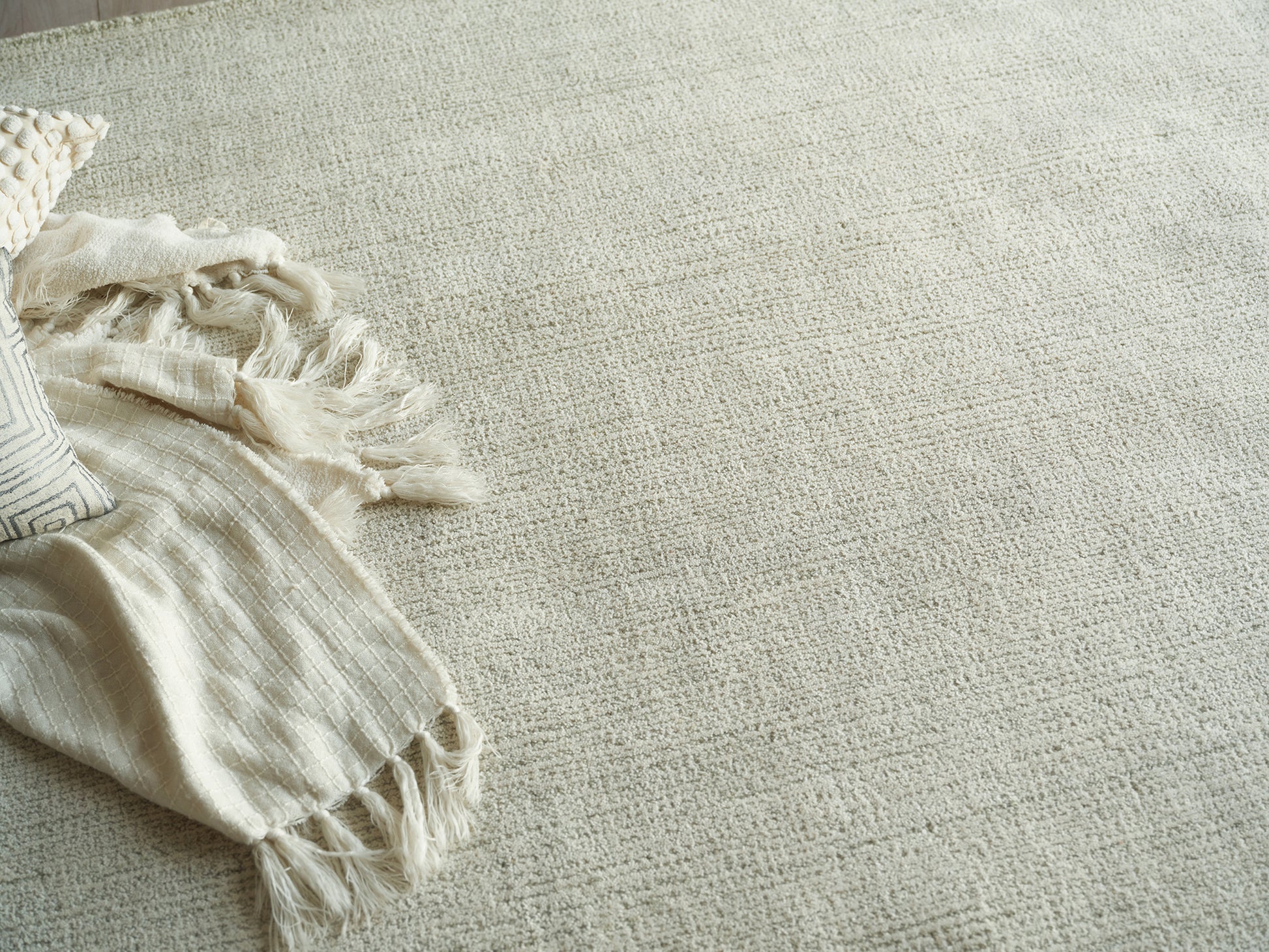 Beige woven area rug with subtle texture and fringe; cream knit throw blanket and striped pillow on the left.