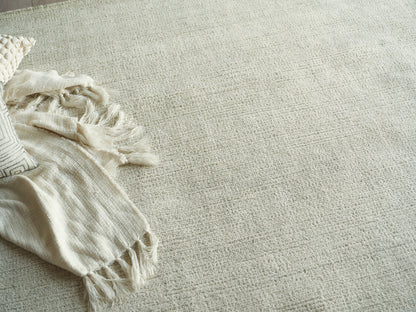 Cream woven rug with a fringed beige throw blanket and a striped pillow on the left, cozy neutral living room look.