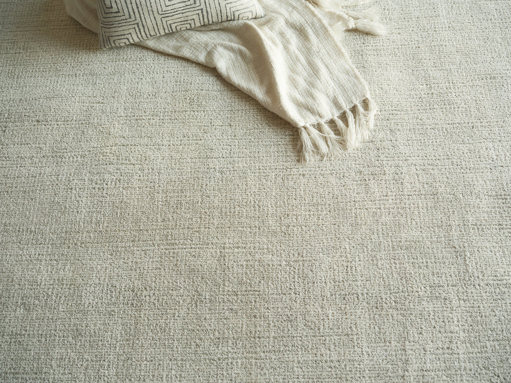 Beige textured rug with cream fringed throw and geometric-patterned pillow.