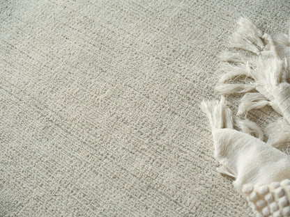 Beige textured area rug with a cream fringed throw draped along its edge.