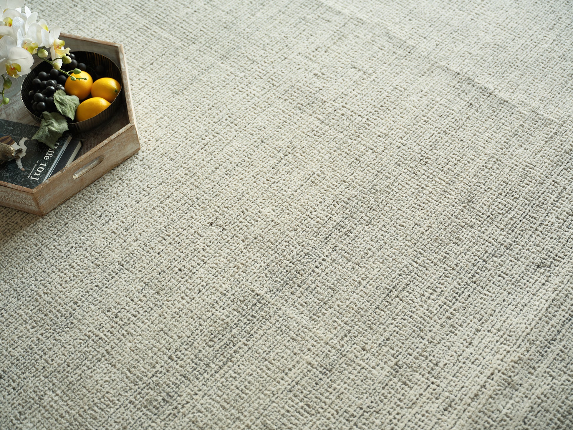 Beige textured rug with a rustic wooden crate holding yellow lemons, black grapes, and white orchids on the left.