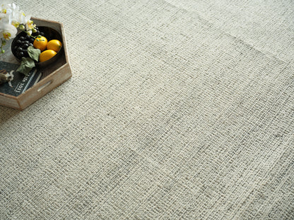 Beige textured woven rug with a top-left wooden crate holding lemons, dark grapes, green leaves and white orchids.