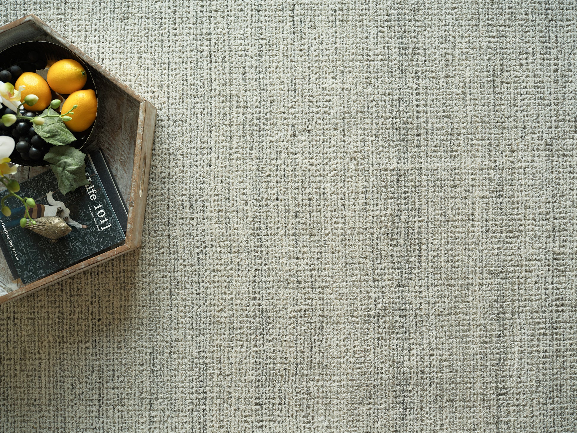 Wooden crate with yellow citrus, dark grapes, greenery, a small bird figurine and a book on a beige textured rug.