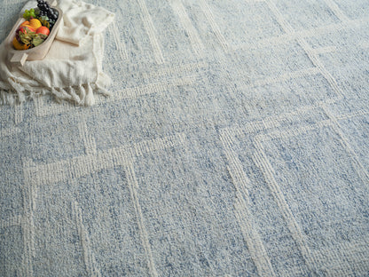 Light blue textured rug with a beige geometric grid; small wooden basket of colorful fruits on a fringed cloth.