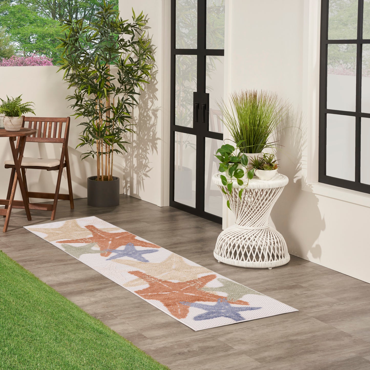 Long outdoor runner rug with orange and blue starfish pattern on beige, beside potted plants and glass doors.