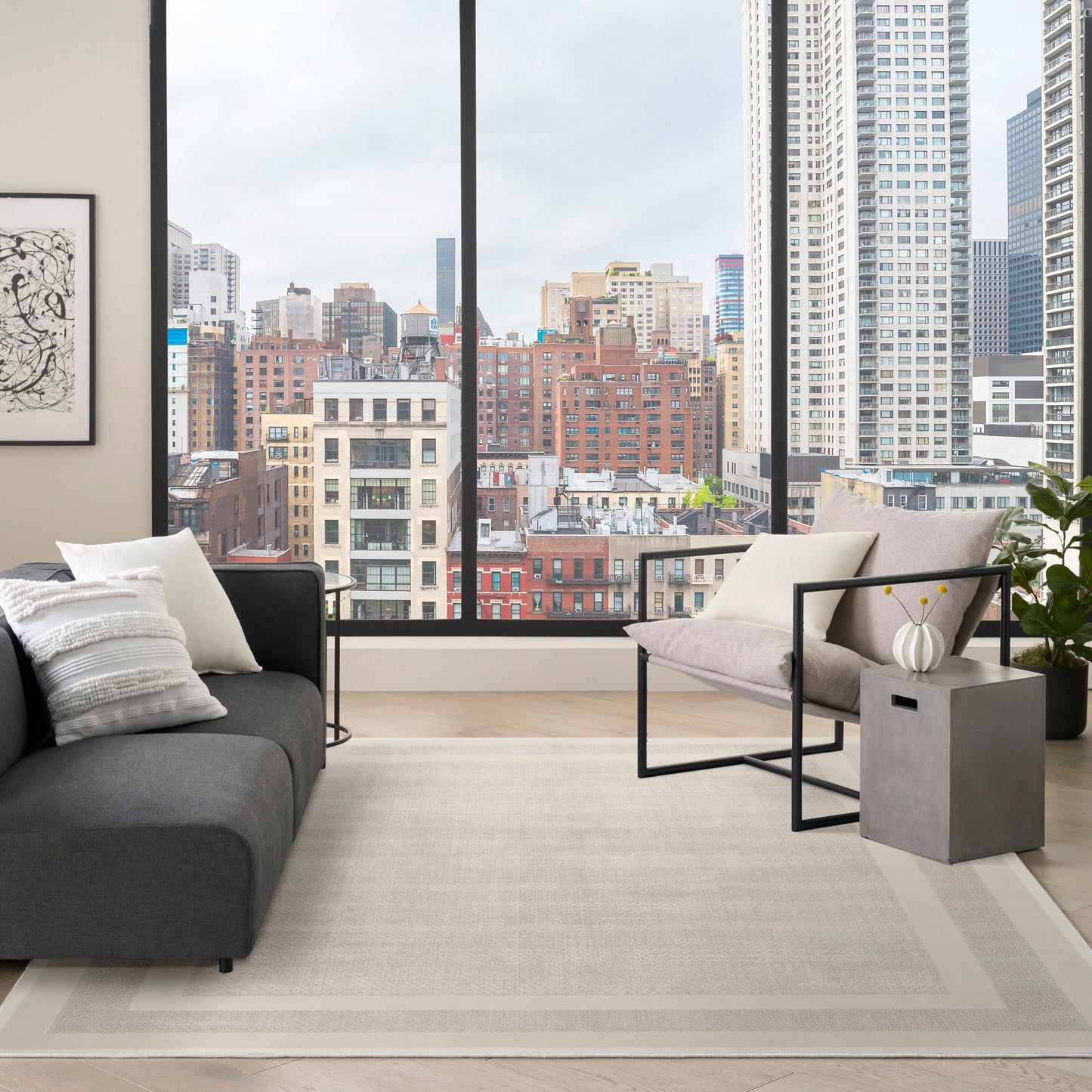 Modern living room with city skyline view, dark gray sofa, light gray lounge chair, beige rug (8' x 10' rug), black metal accents and plant.