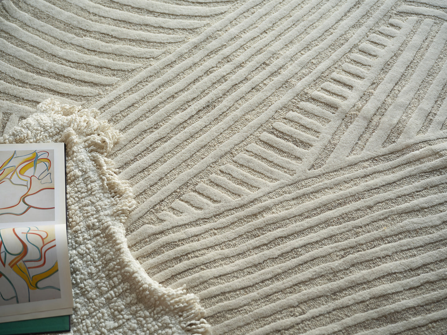 Cream textured area rug with raised linear patterns; fringed beige throw and colorful abstract art magazine nearby.