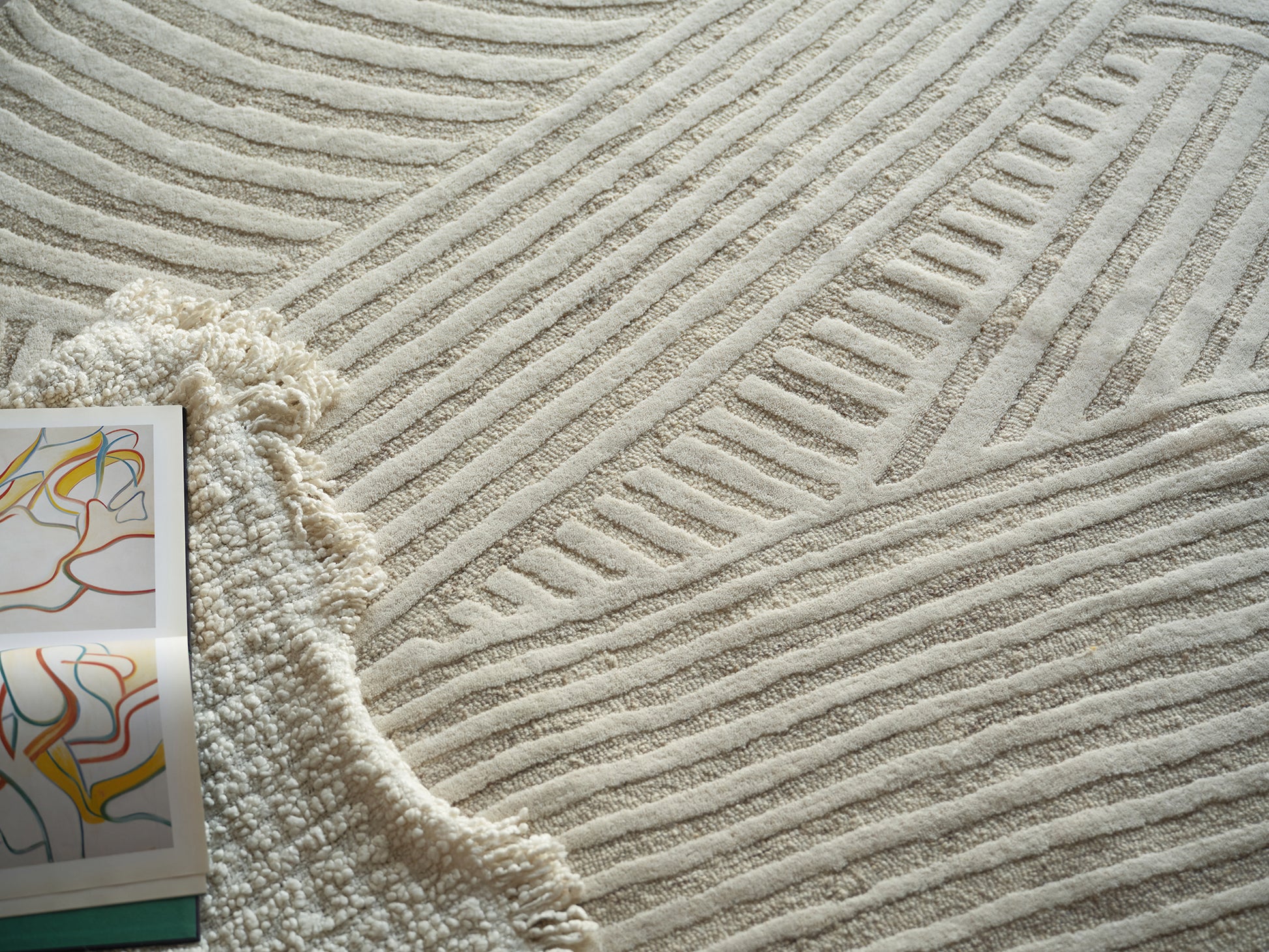 Cream textured area rug with raised linear patterns; fringed beige throw and colorful abstract art magazine nearby.