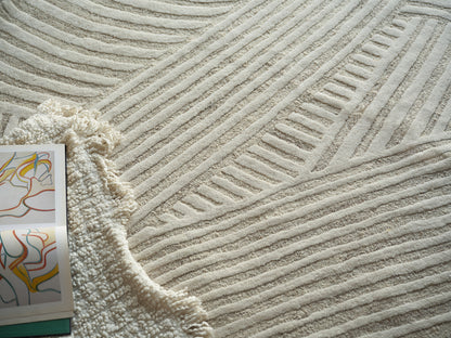 Cream textured area rug with raised linear patterns; fringed beige throw and colorful abstract art magazine nearby.