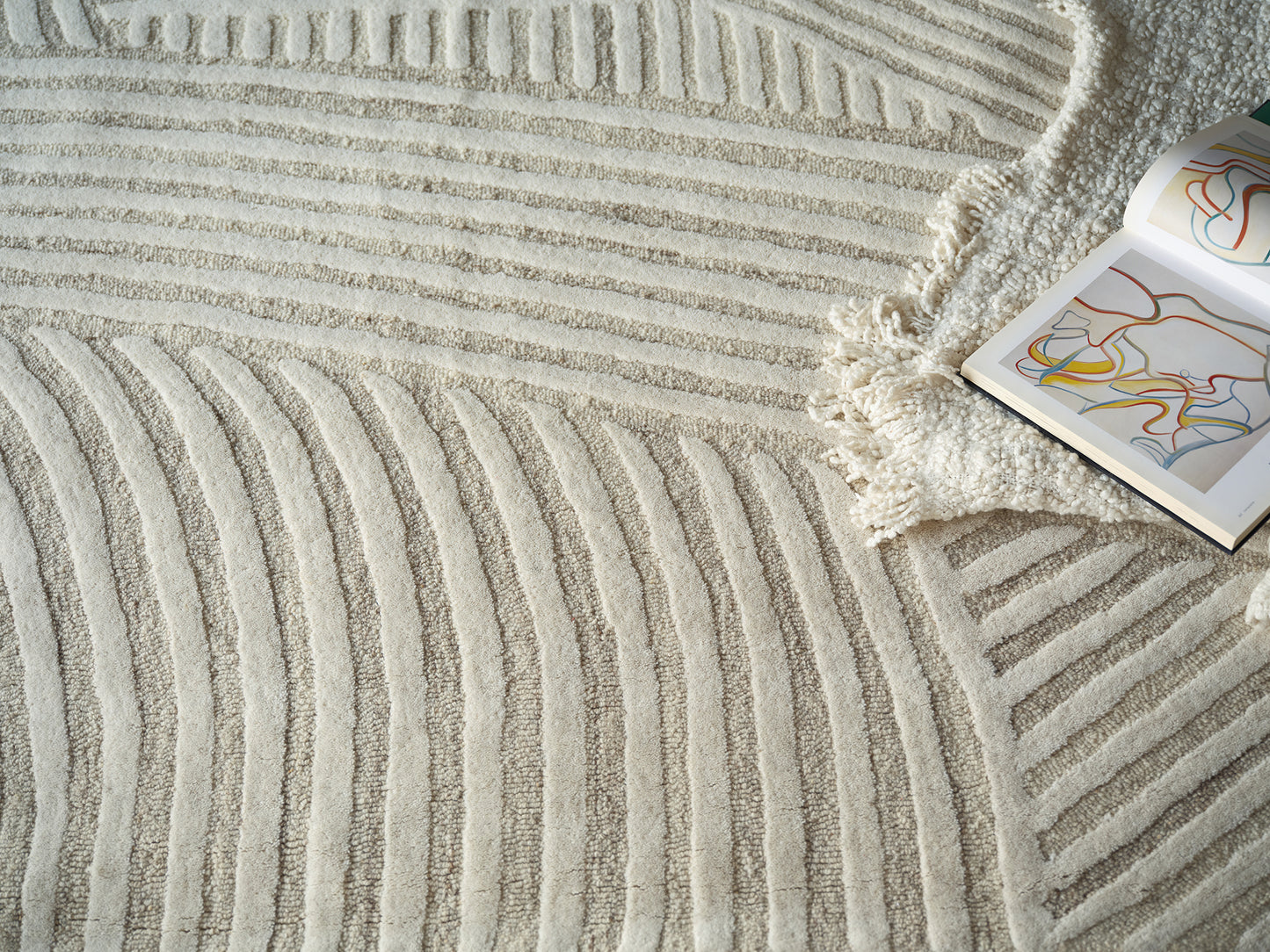 Cream shag rug with raised curved stripe pattern and fringe, beside an open art book with colorful abstract lines.